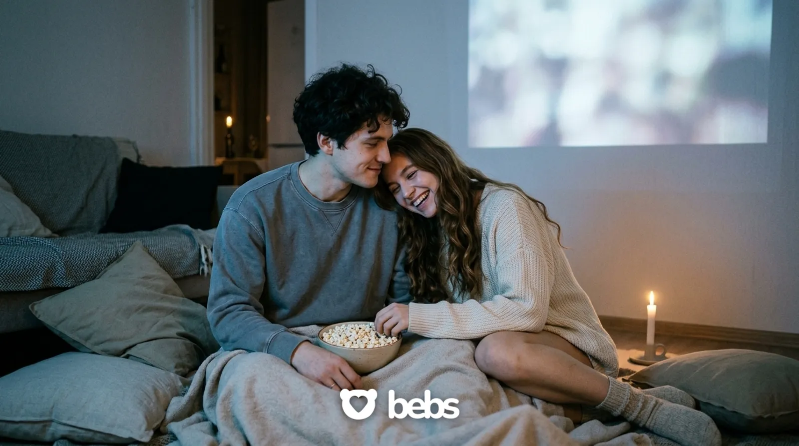 Couple cuddled in blankets watching a movie by projector light at home, cozy third date ideas for a night in